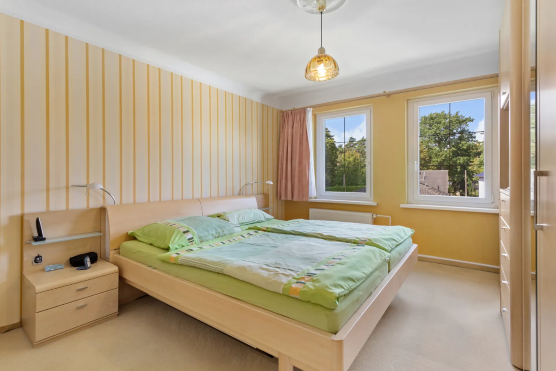 Schlafzimmer mit Doppelbett, Nachttisch, Kleiderschrank und drei Fenstern mit Blick ins Grüne.