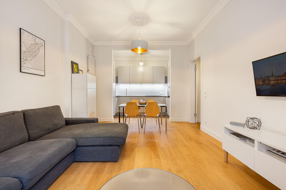 Modernes Wohnzimmer mit grauem Sofa, Esstisch, TV und offener Küche, Holzboden.
