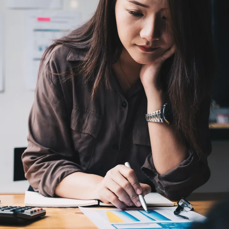 Frau sitzt an einem Schreibtisch mit Notizbuch, Diagramm, Taschenrechner und Pflanze.