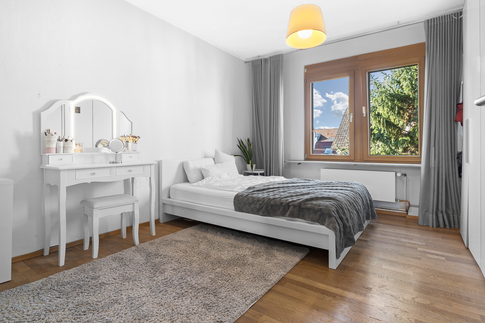 Helles Schlafzimmer mit Doppelbett, Schminktisch, großem Fenster und Vorhängen auf Holzfußboden.