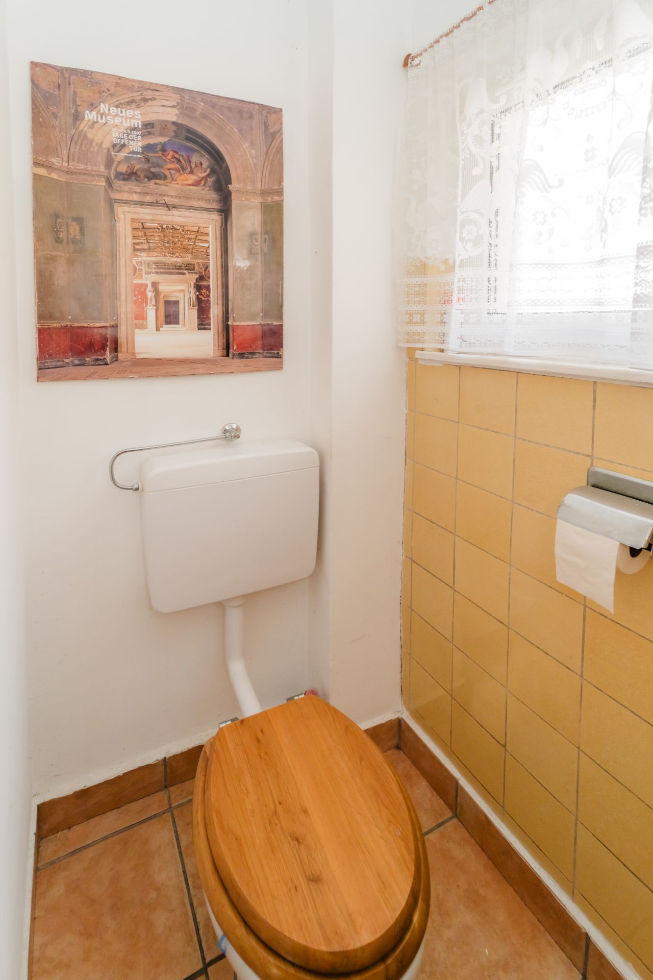 Kleines WC mit Holzklo, gelben Fliesen, Fenster mit Gardine und Bild an der Wand.