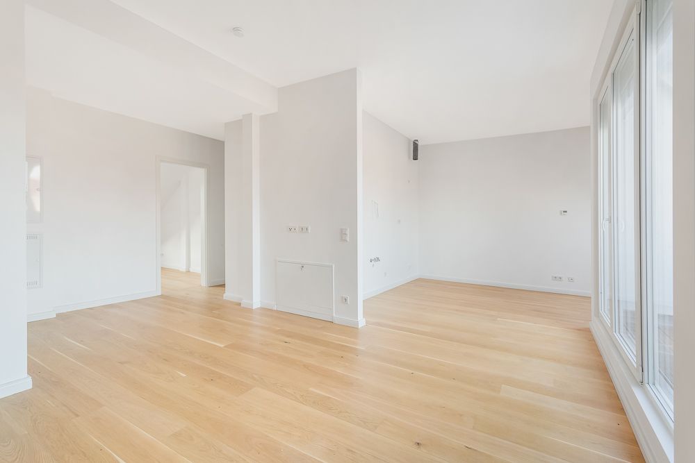 Heller, leerer Raum mit Holzboden und weißen Wänden, große Fenster auf der rechten Seite.