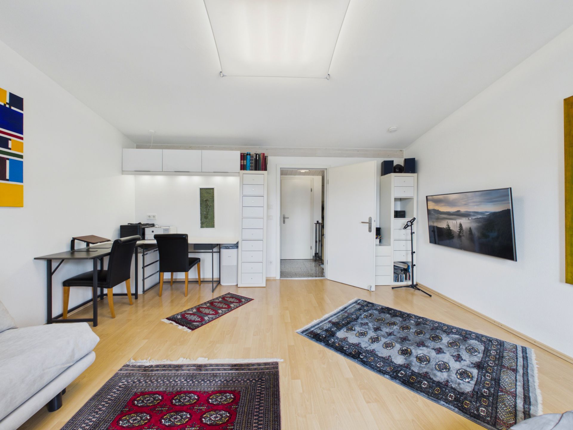 Helles Wohnzimmer mit Parkettboden, Schreibtisch, Teppichen und Wandfernseher.