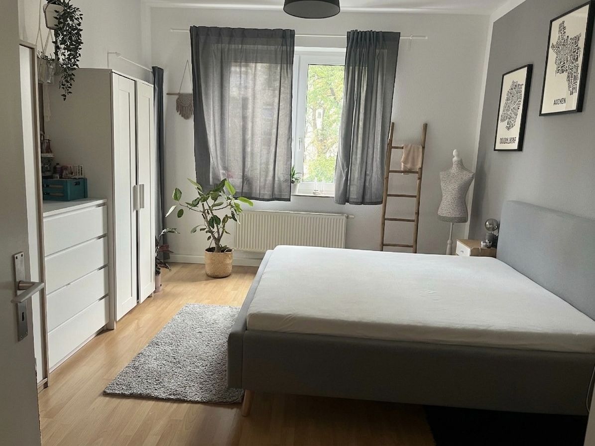Helles Schlafzimmer mit grauem Bett, Schrank, Kommode, Pflanze und Fenster mit Vorhängen.