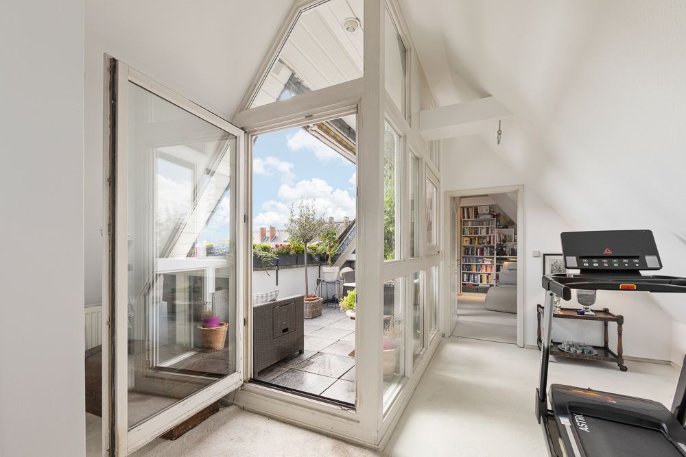 Heller Raum mit großen Fenstern, Zugang zur Terrasse, Laufband und Blick ins Wohnzimmer mit Bücherregal.