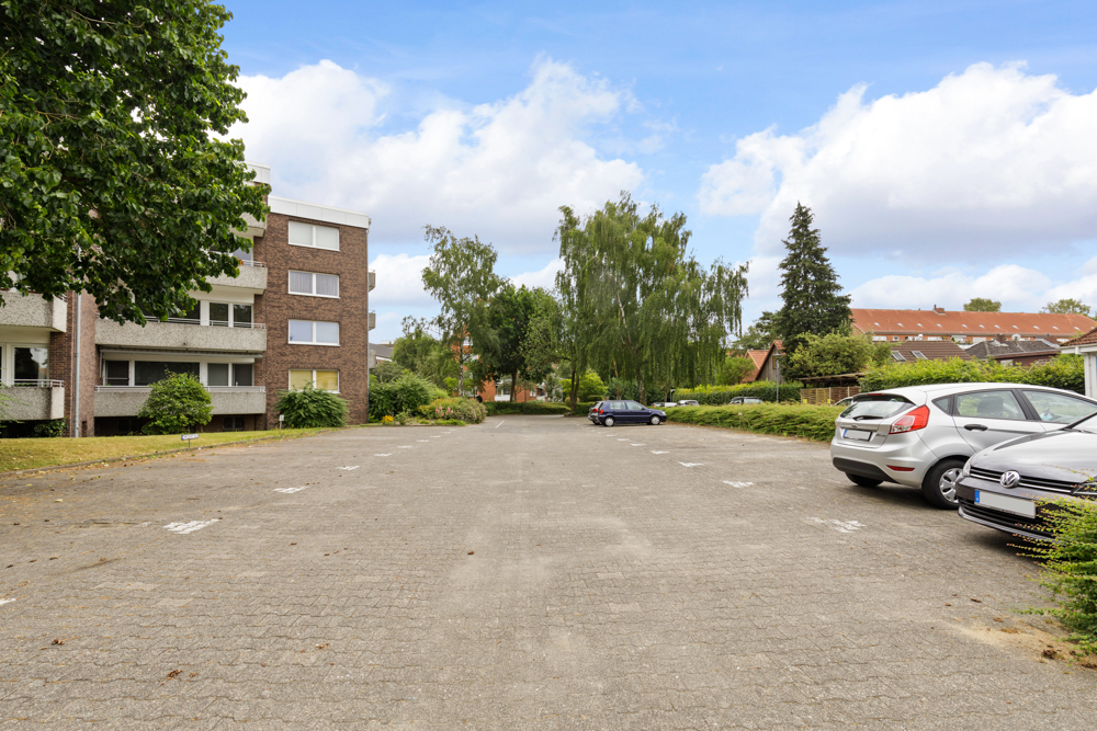 Wohngebäude mit angrenzendem Parkplatz, mehreren Autos und Bäumen im Hintergrund.