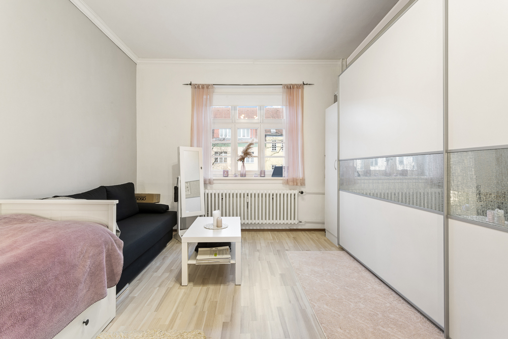 Helles Zimmer mit Bett, Sofa, Couchtisch, großem Schrank und Fenster mit rosa Vorhängen.