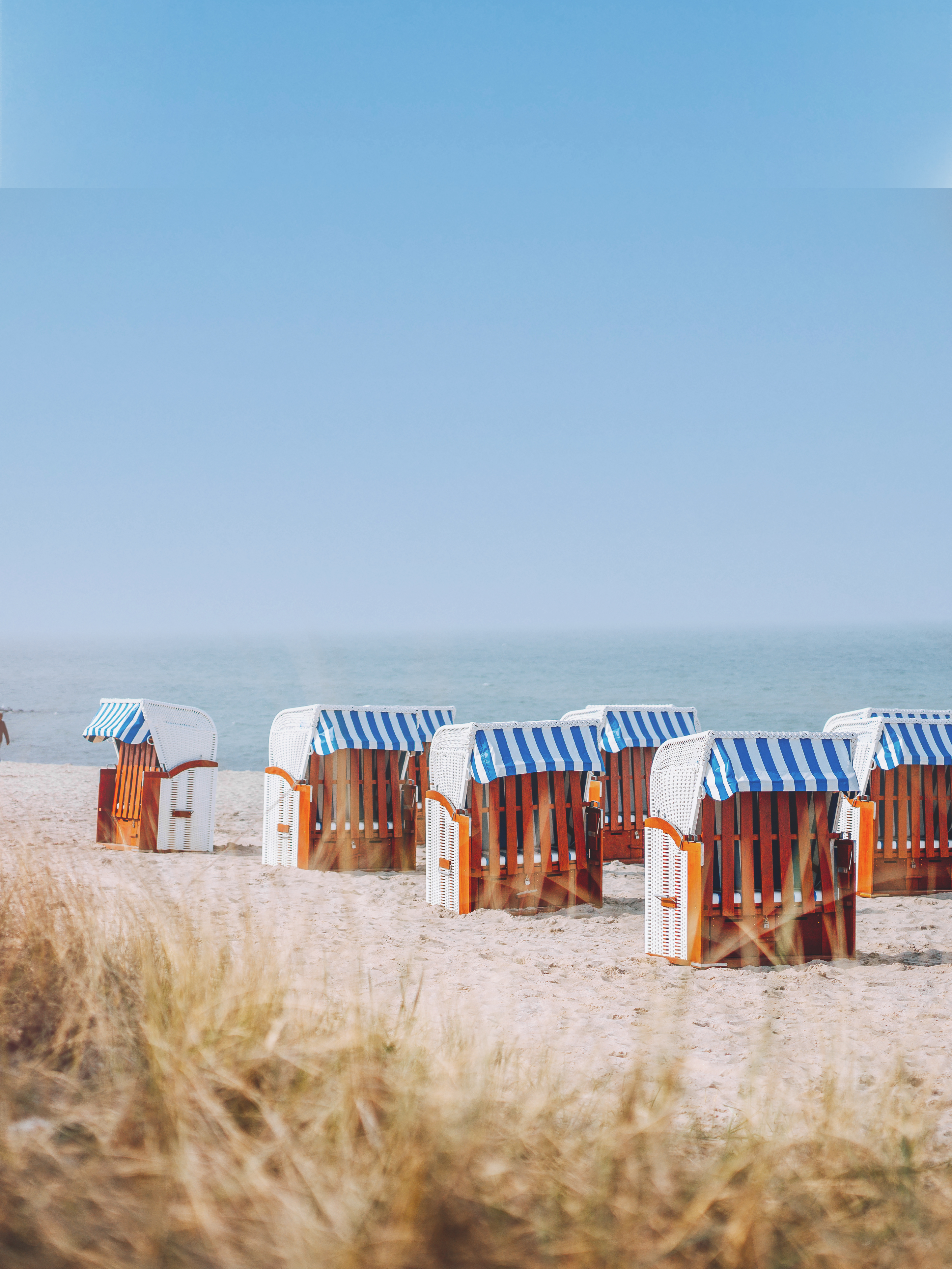 Mehrere Strandkörbe mit blau-weiß gestreiften Dächern stehen am Sandstrand vor dem Meer.