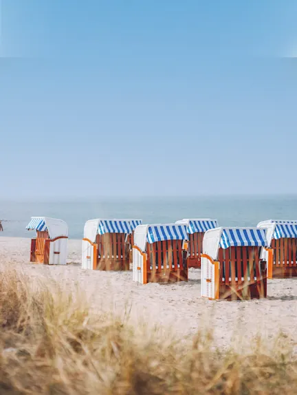 Mehrere Strandkörbe mit blau-weiß gestreiften Dächern stehen am Sandstrand vor dem Meer.