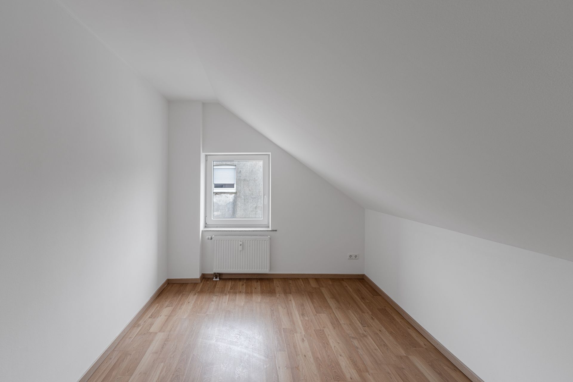 Leerer Raum mit Dachschräge, weiß gestrichenen Wänden und Holzfußboden, Fenster und Heizung an der Stirnwand.