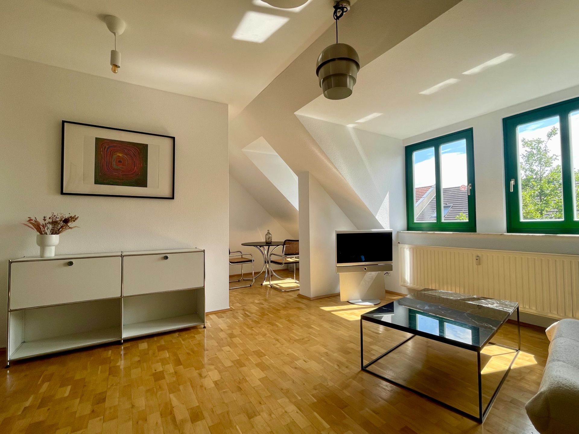 Helles Wohnzimmer mit Holzboden, Couchtisch, Sideboard, Fernseher und zwei Fenstern mit grünen Rahmen.