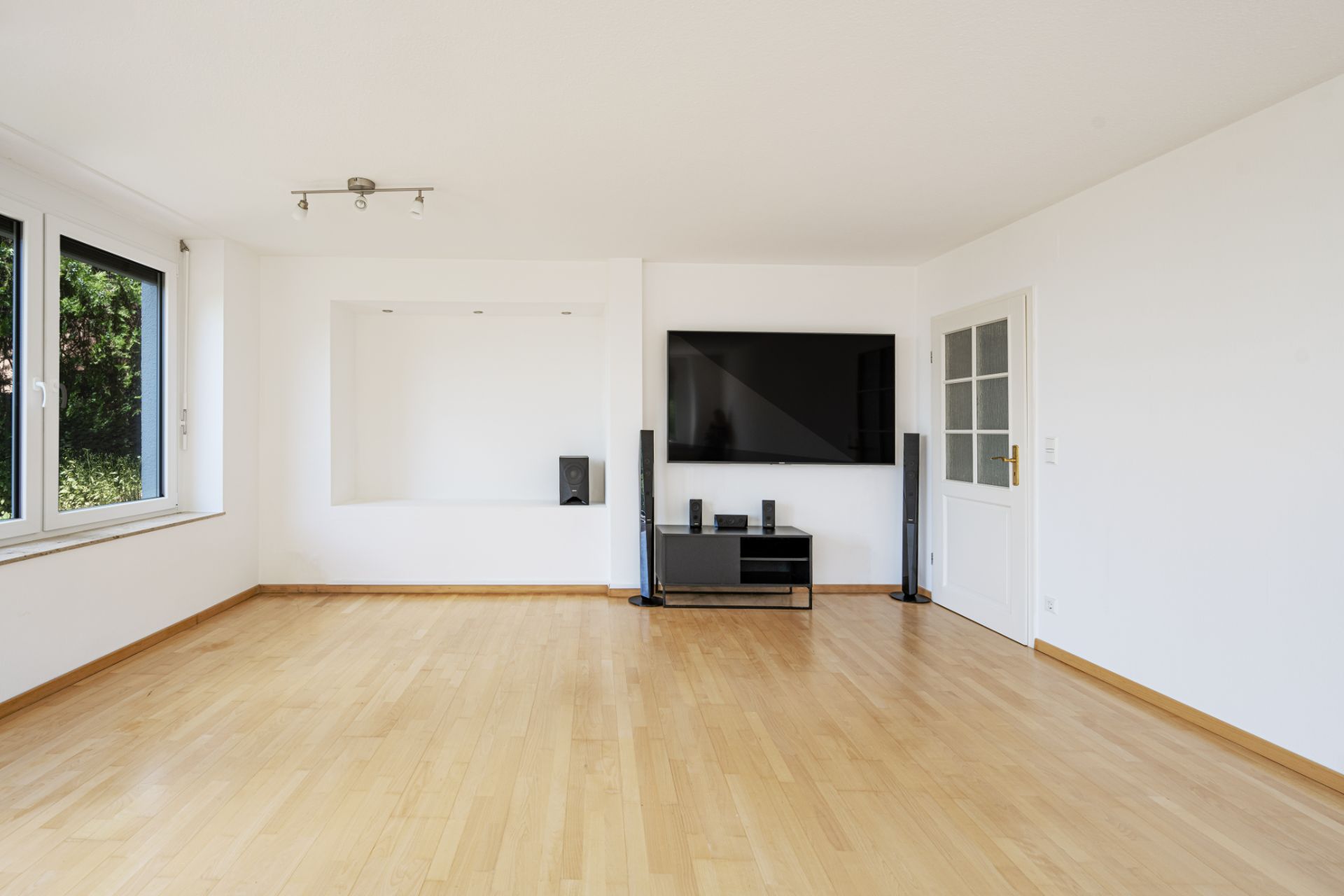 Helles Wohnzimmer mit Holzboden, großem Fenster, Fernseher und Glastür.