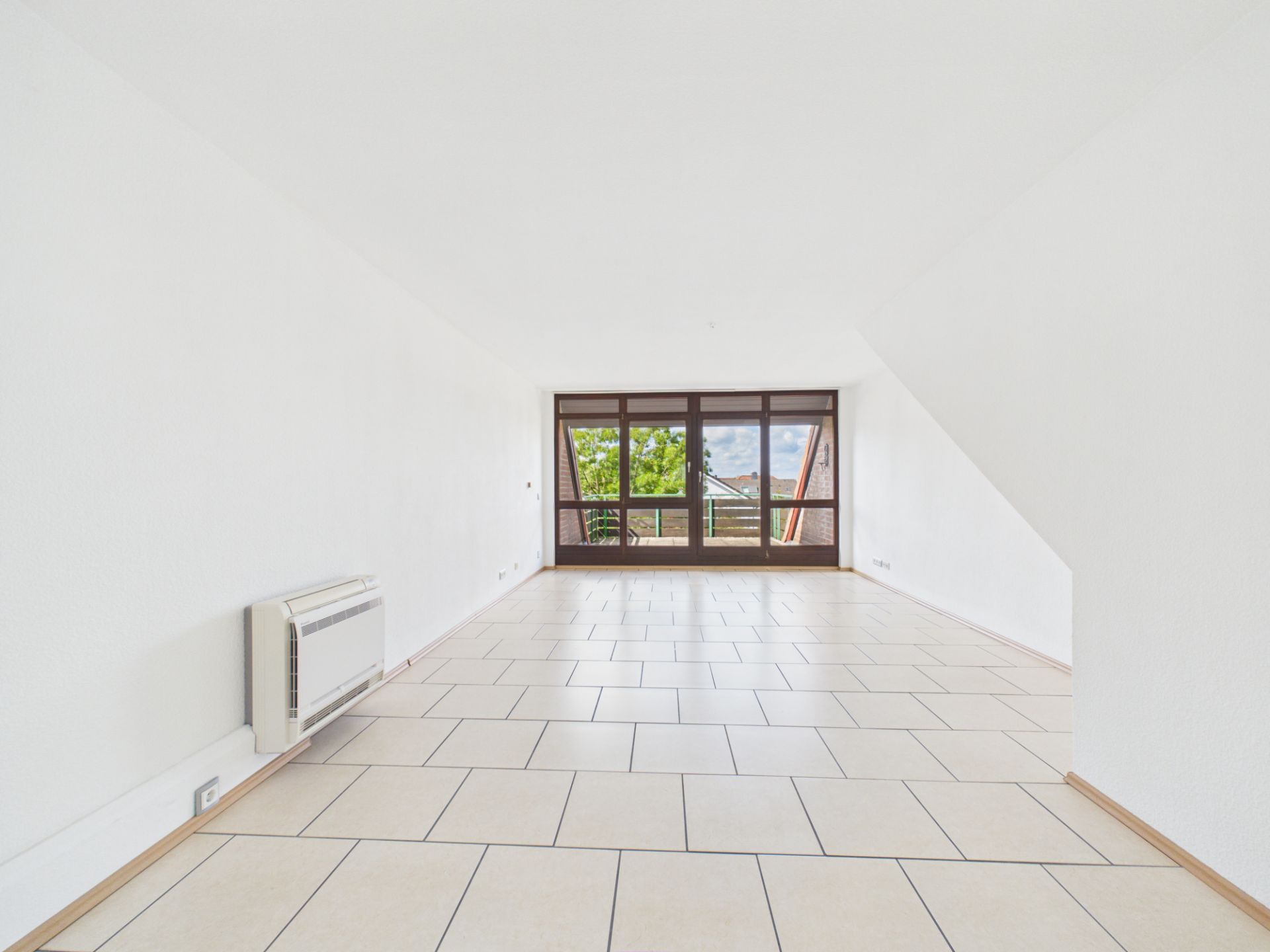 Helles, leeres Zimmer mit Fliesenboden und großen Fenstern mit Blick ins Grüne.