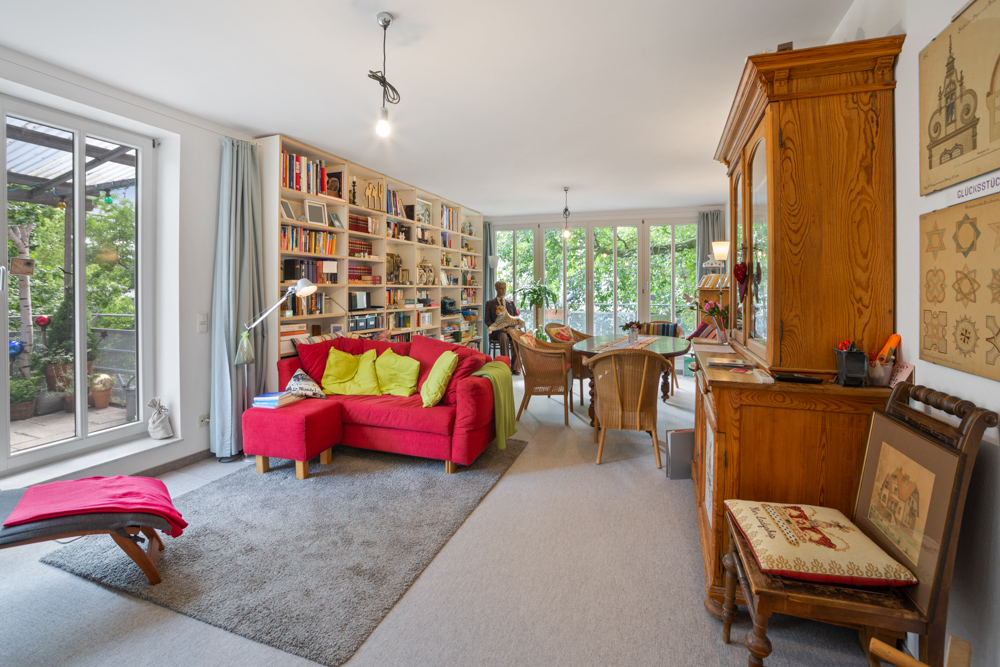 Helles Wohnzimmer mit rotem Sofa, Bücherregal, Holzmöbeln und Blick auf eine Terrasse mit Pflanzen.
