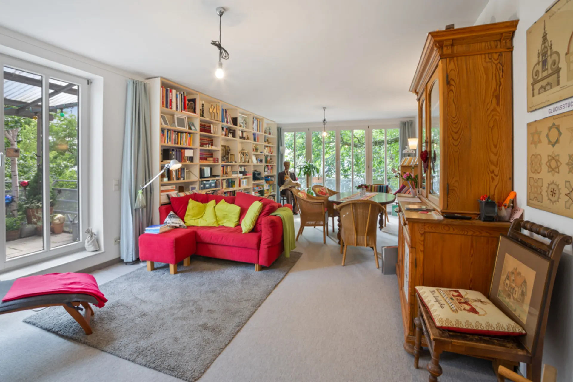 Helles Wohnzimmer mit rotem Sofa, Bücherregal, Holzmöbeln und Blick auf eine Terrasse mit Pflanzen.