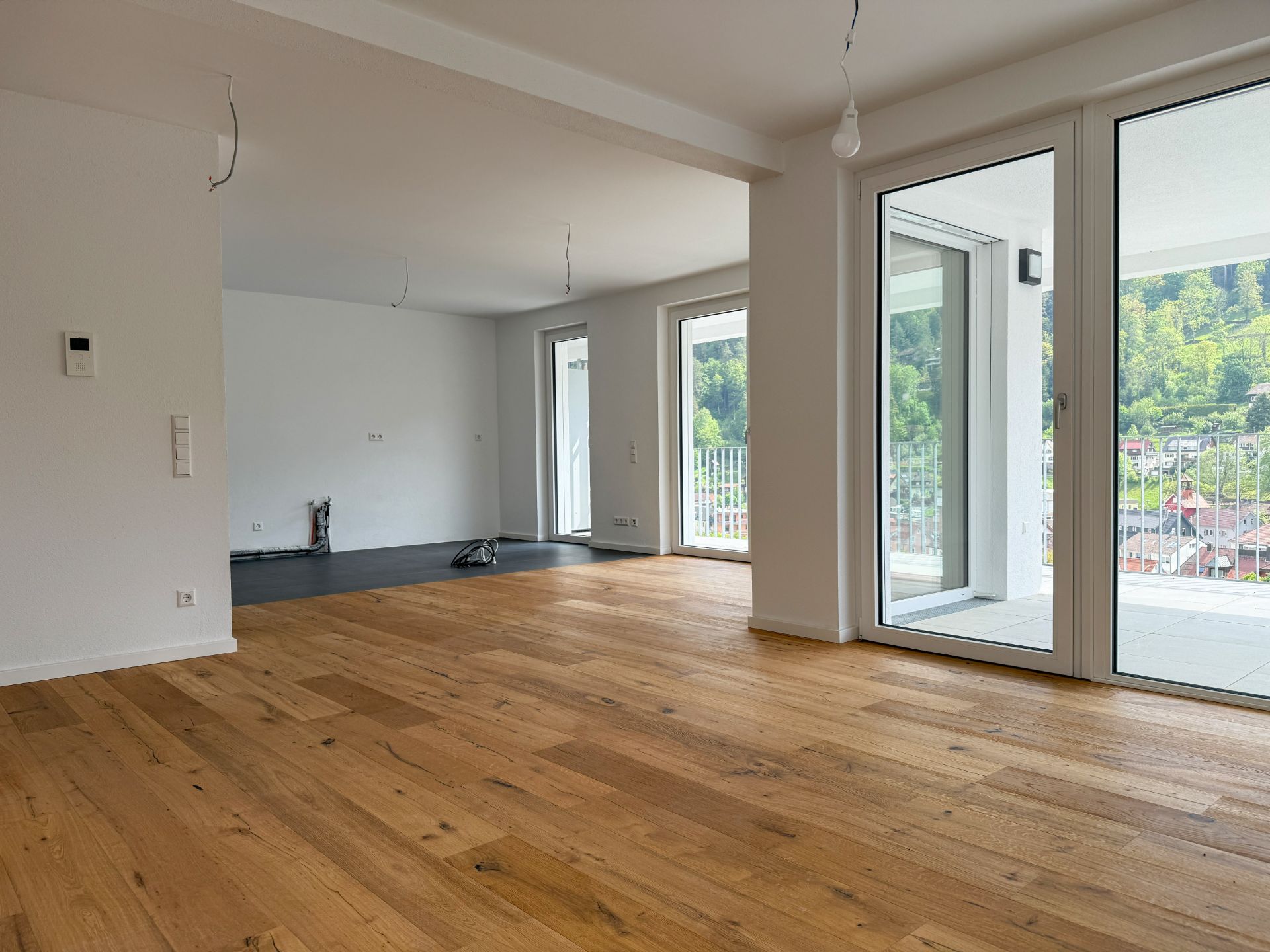 Leeres, helles Wohnzimmer mit Holzboden und großen Fenstern zum Balkon.