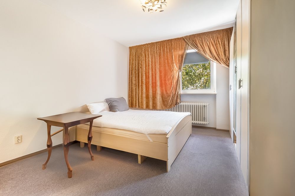 Helles Schlafzimmer mit Doppelbett, Tisch, Kleiderschrank und Fenster mit braunen Vorhängen.