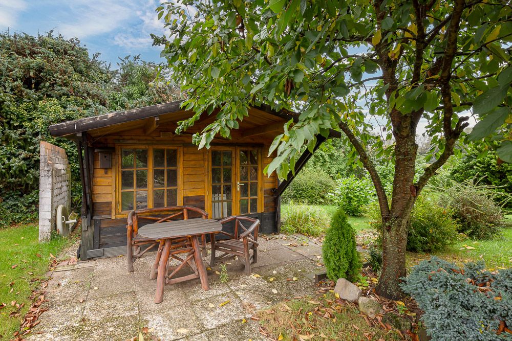 Holzgartenhaus mit Terrasse, Holztisch und Stühlen, von Bäumen und Sträuchern umgeben.