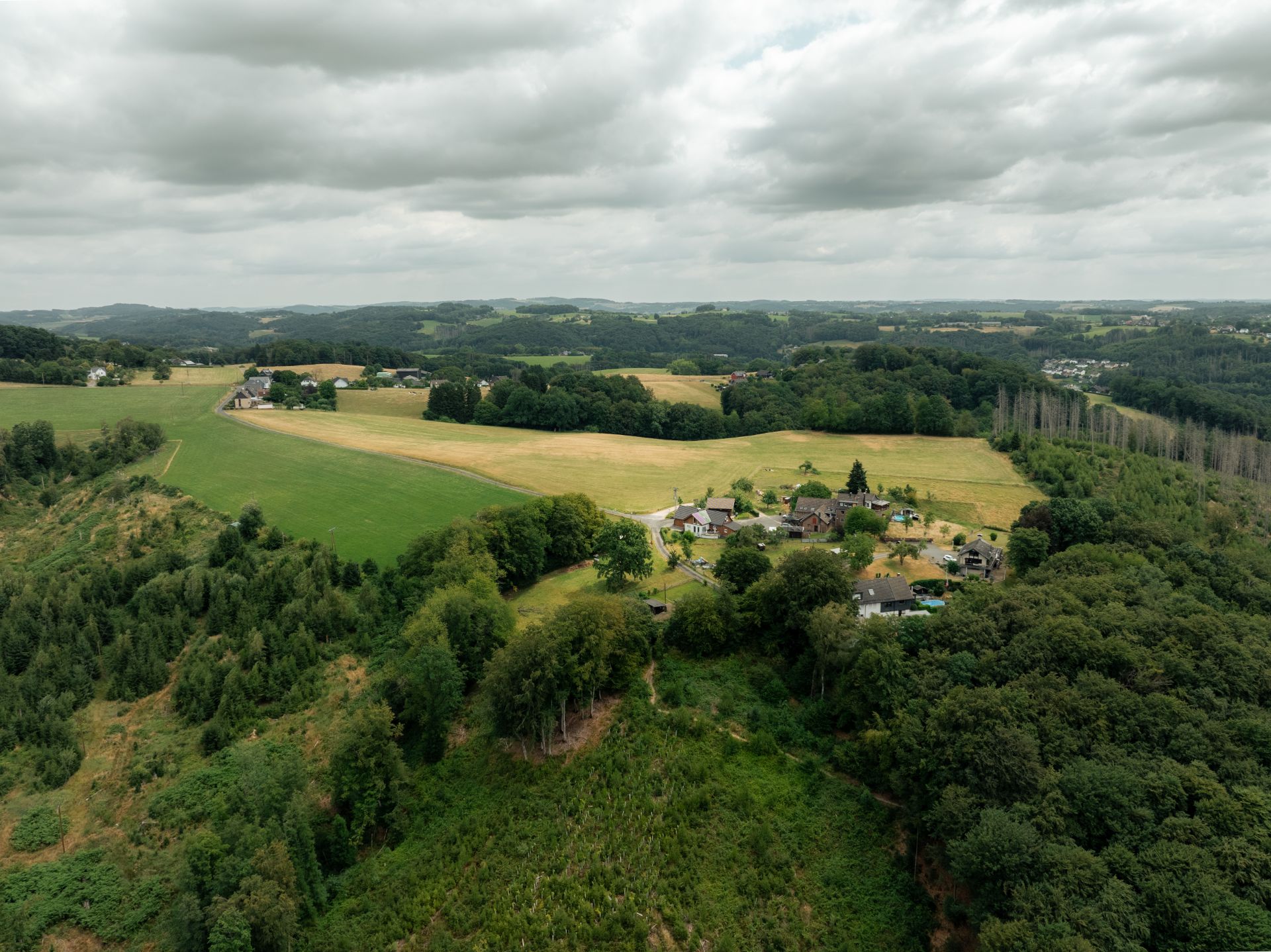 Luftaufnahme eines kleinen Dorfes umgeben von Wäldern und Feldern unter bewölktem Himmel.
