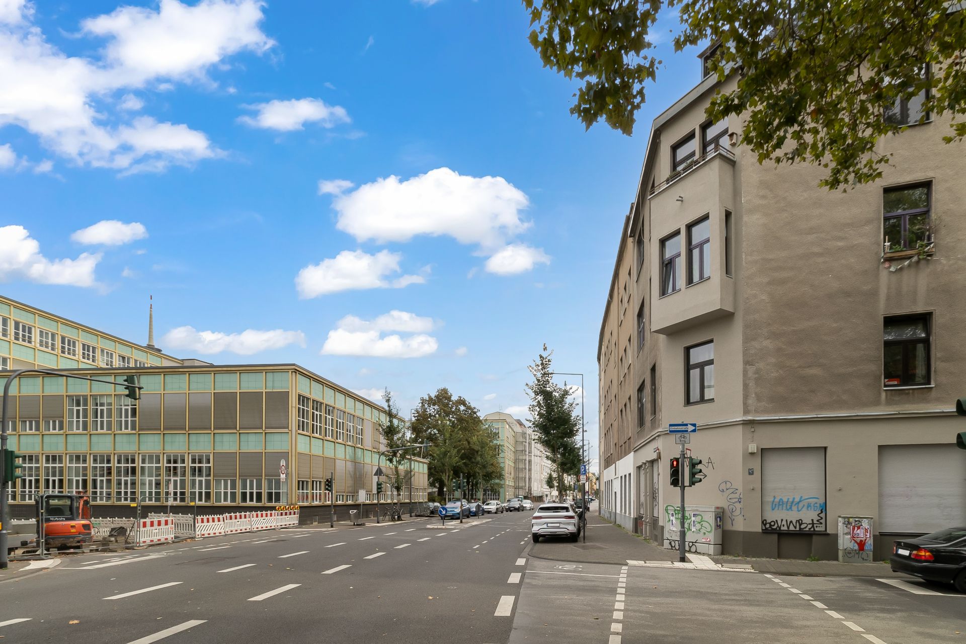 Breite Straße mit mehrstöckigen Wohnhäusern, parkenden Autos und Bäumen am Rand.