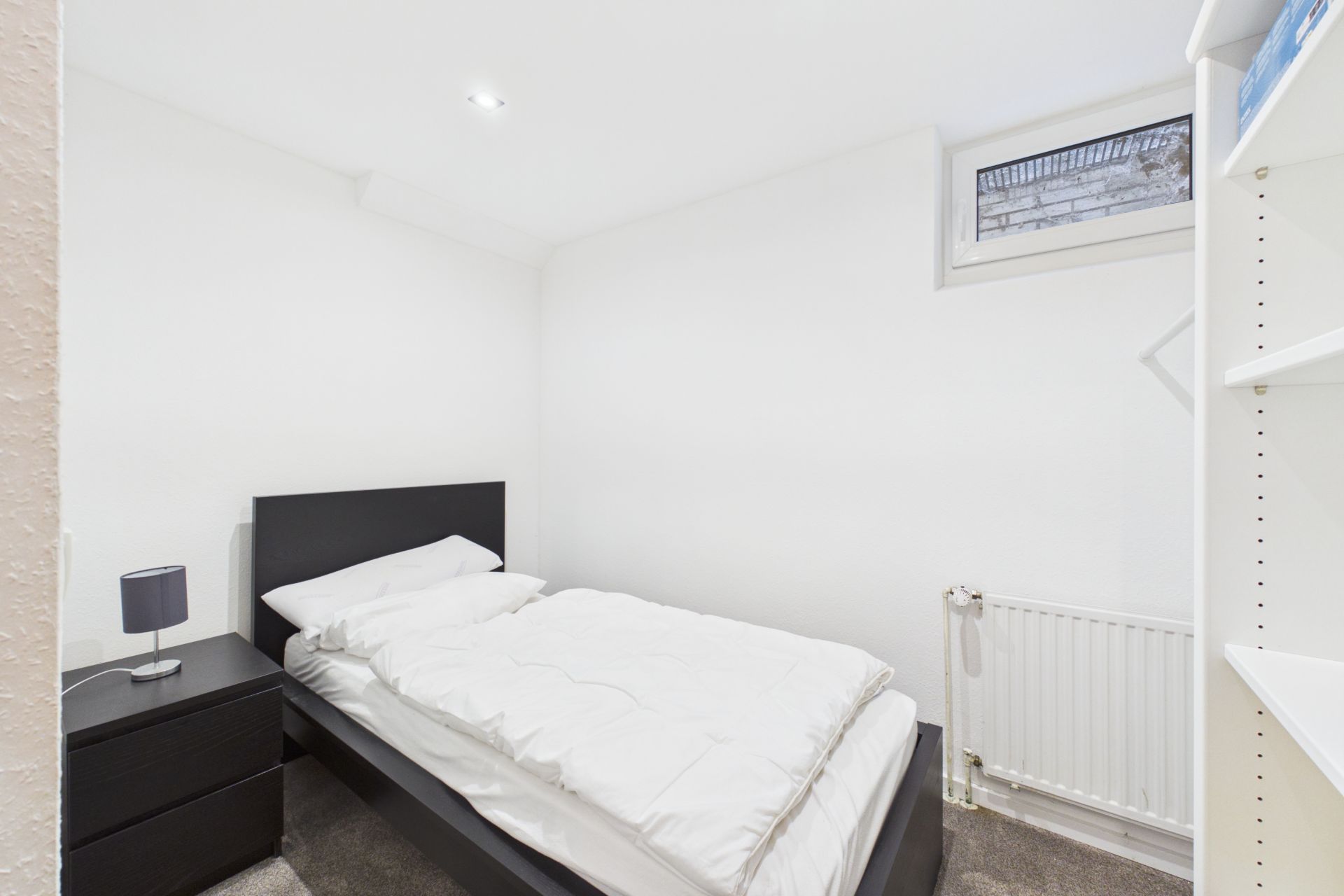 Kleines Schlafzimmer mit Einzelbett, Nachttisch, Regal, Heizkörper und Fenster hoch an der Wand.