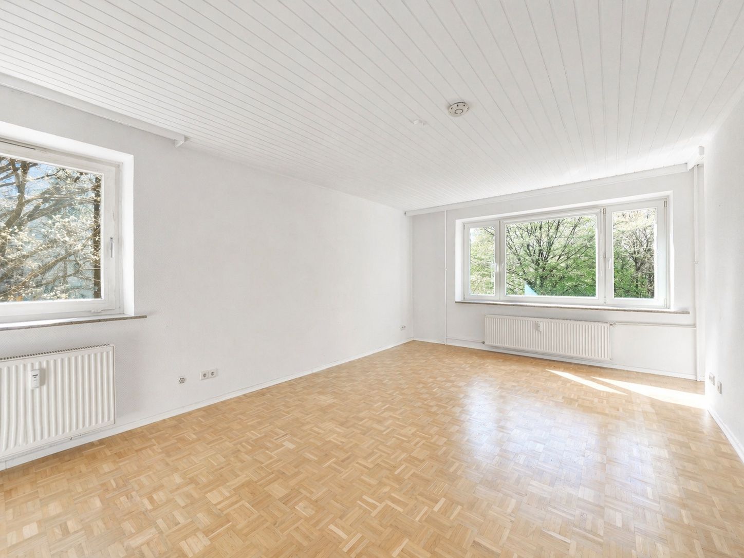 Heller, leerer Raum mit Parkettboden, weißen Wänden und großen Fenstern mit Blick ins Grüne.