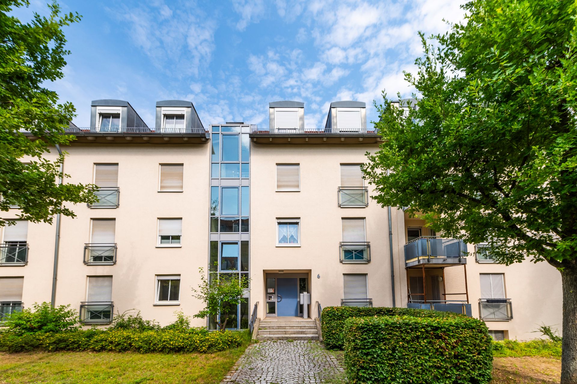 Mehrfamilienhaus mit vier Etagen, Balkonen und Eingangsbereich, umgeben von Bäumen und Grünflächen.