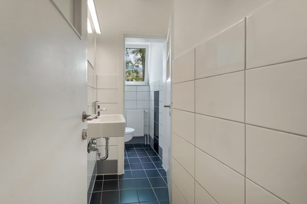 Schmales Badezimmer mit kleinem Waschbecken, Toilette, weißen Fliesen und Fenster.