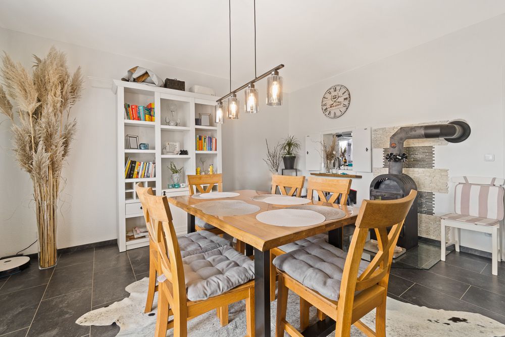 Essbereich mit Holztisch, sechs Stühlen, Bücherregal und Kaminofen.
