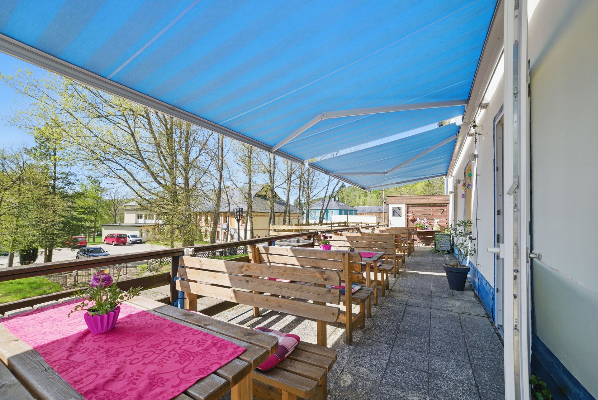 Terrasse mit Holzbänken, Tischen mit rosa Decken und blauen Markisen, Blumen auf den Tischen.