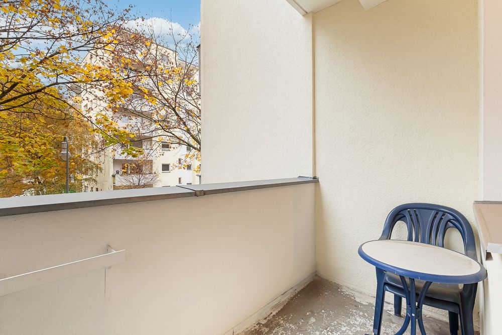 Balkon mit zwei Kunststoffstühlen und kleinem Tisch, daneben herbstliche Bäume und Wohngebäude.