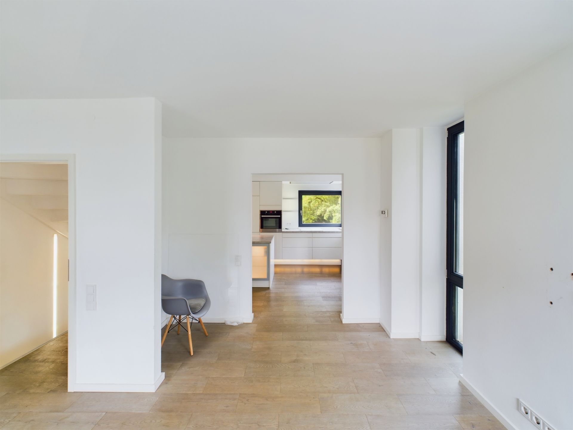 Heller Raum mit Holzfußboden, grauem Stuhl und Blick in eine moderne Küche.