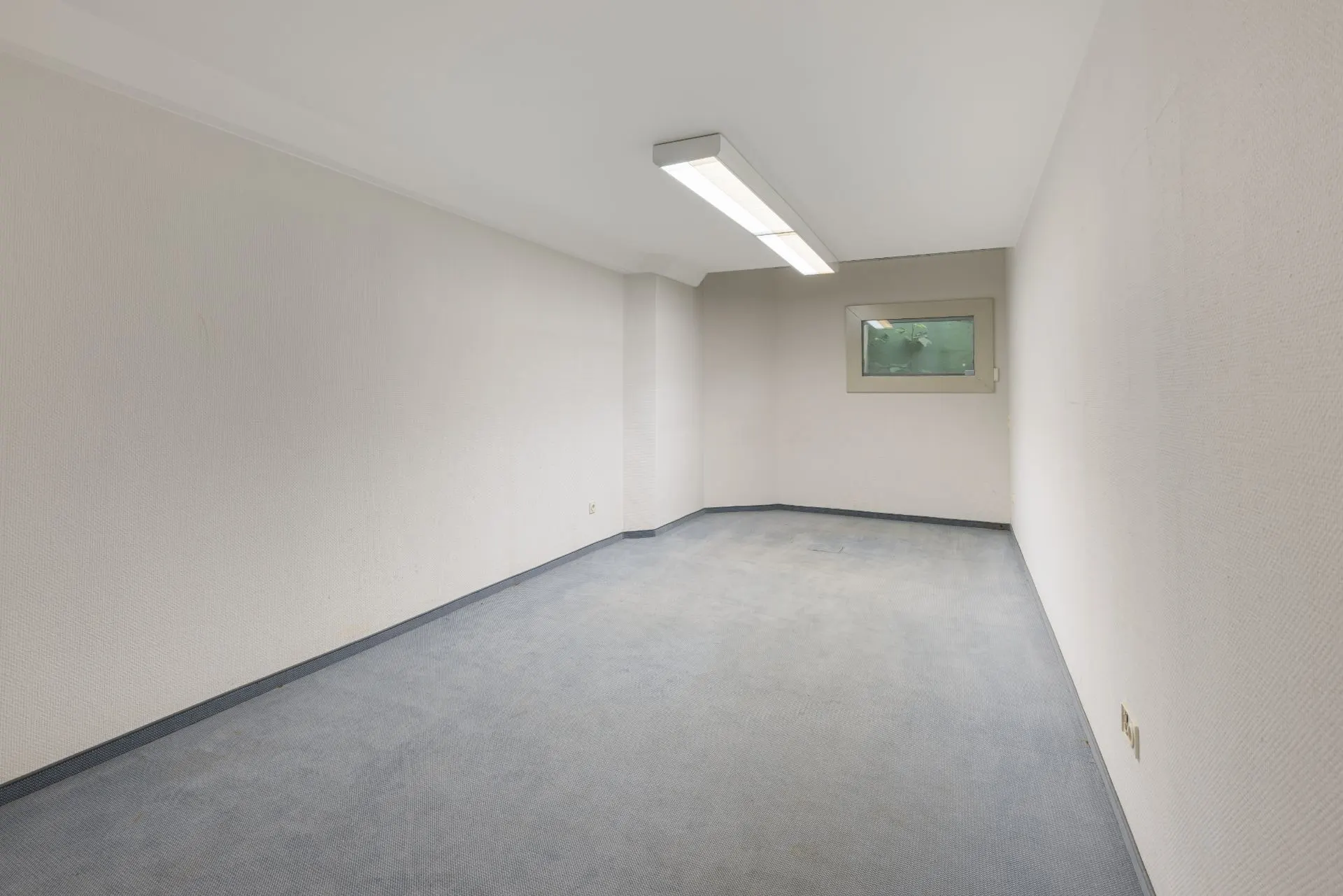 Leerer Raum mit Teppichboden, weißen Wänden, Deckenleuchte und einem kleinen Fenster.