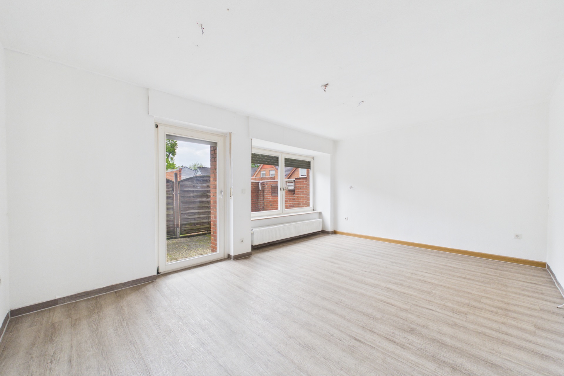 Leeres, helles Wohnzimmer mit großem Fenster, Glastür und Holzfußboden.