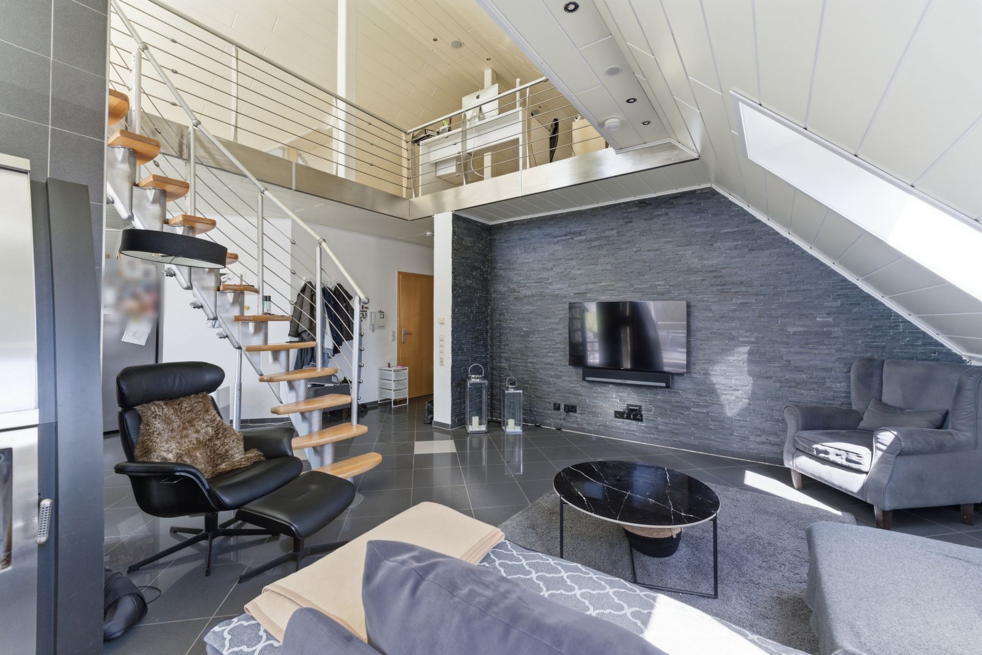 Modernes Wohnzimmer mit offener Treppe, Fernseher an Steinwand und Sitzmöbeln unter Dachschräge.