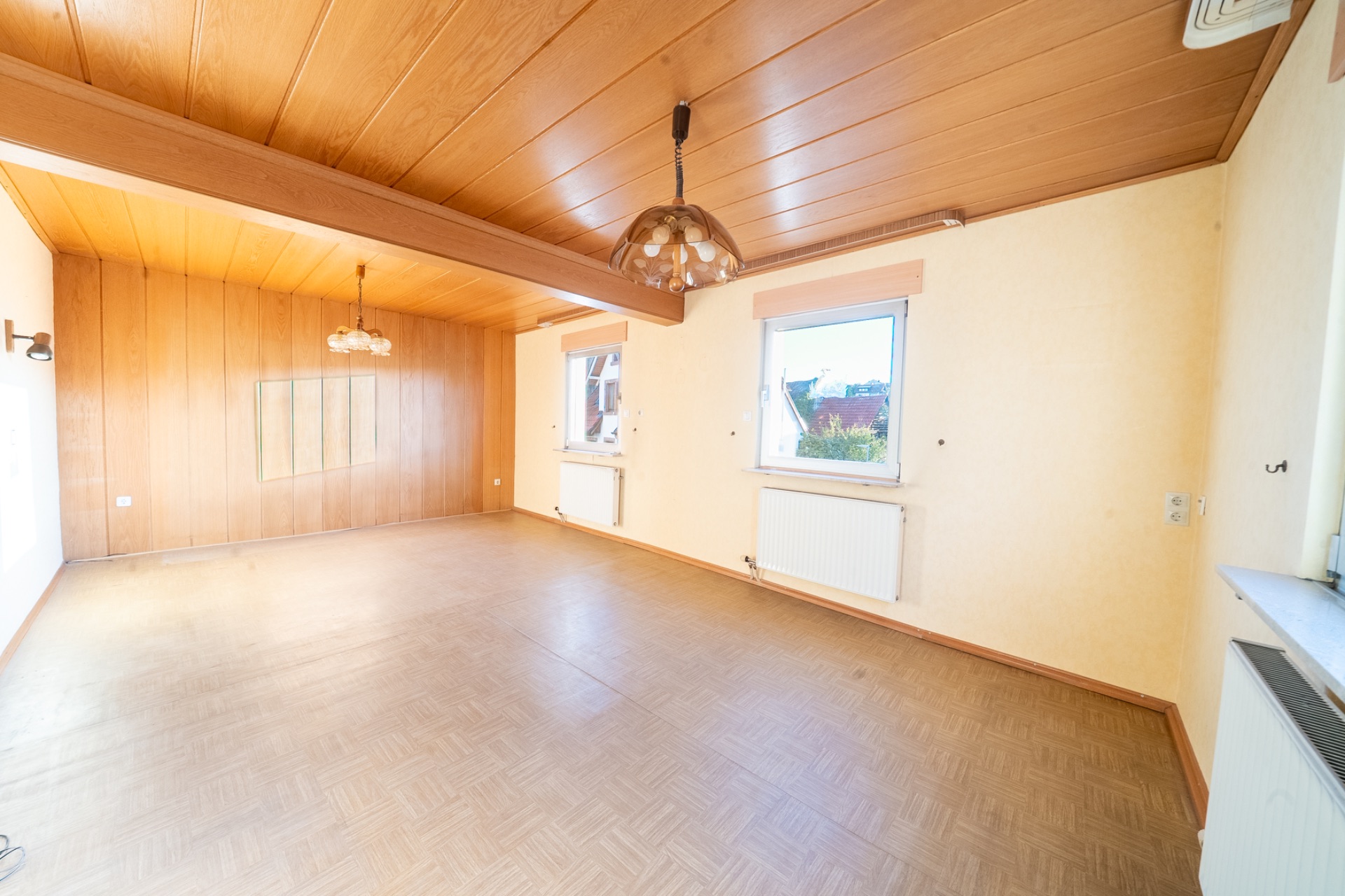 Heller, leerer Raum mit Holzdecke, Holzwandverkleidung, Fenstern und zwei Hängelampen.