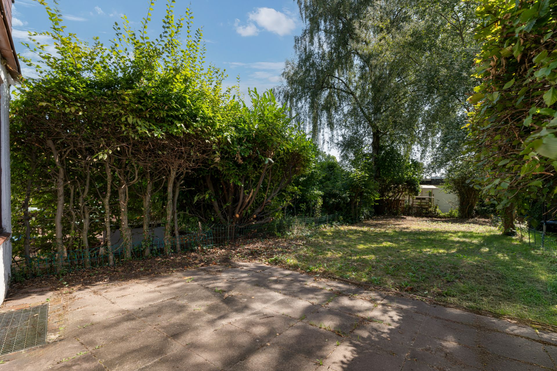 Garten mit Rasenfläche, Bäumen und einer kleinen Terrasse im Schatten.