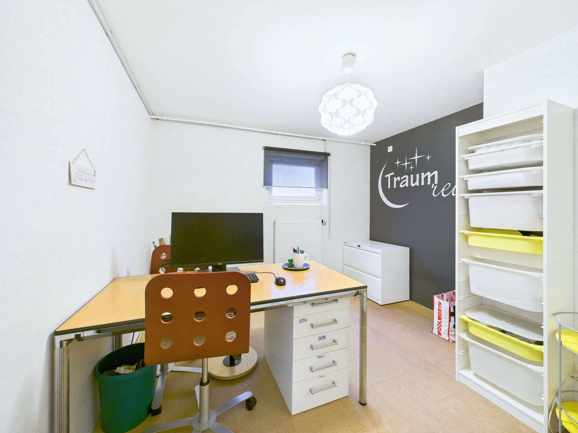 Helles Arbeitszimmer mit Schreibtisch, Stuhl, Regal und Computerbildschirm.