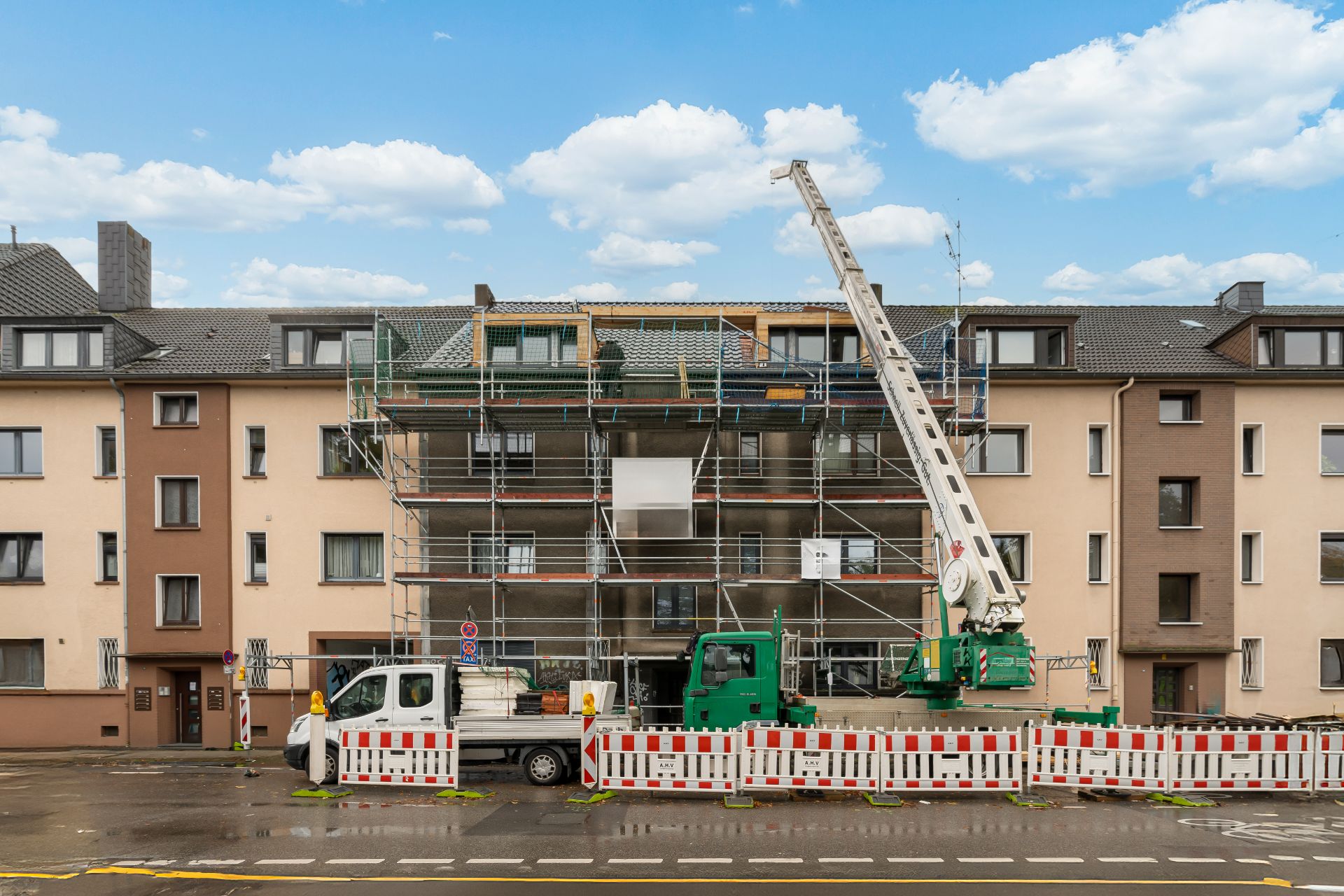 Wohnhaus mit Baugerüst und Kran, davor stehen Absperrungen und Fahrzeuge auf der Straße.