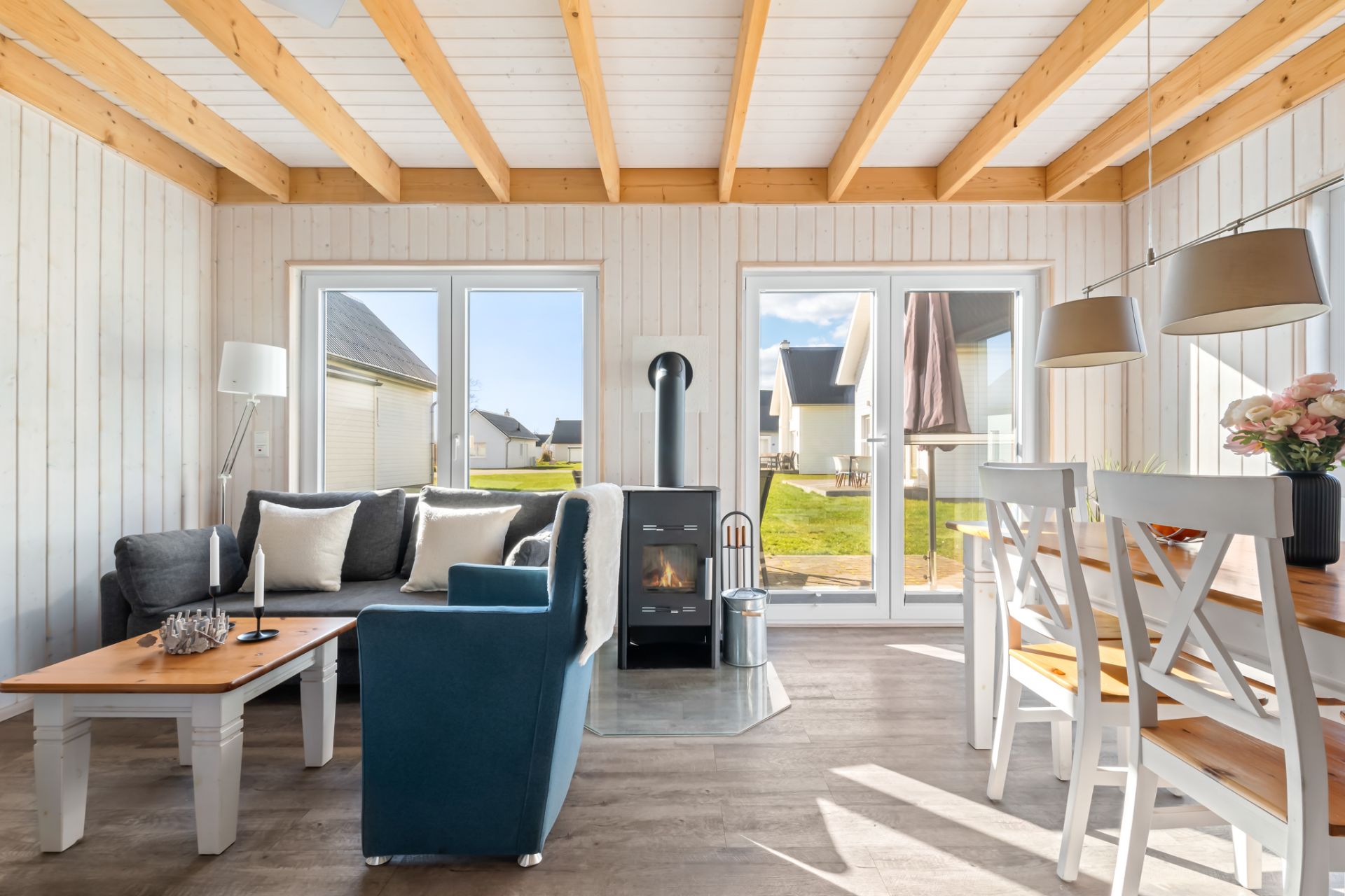 Helles Wohnzimmer mit Sofa, Sessel, Holzofen, Esstisch und großen Fenstern mit Blick nach draußen.