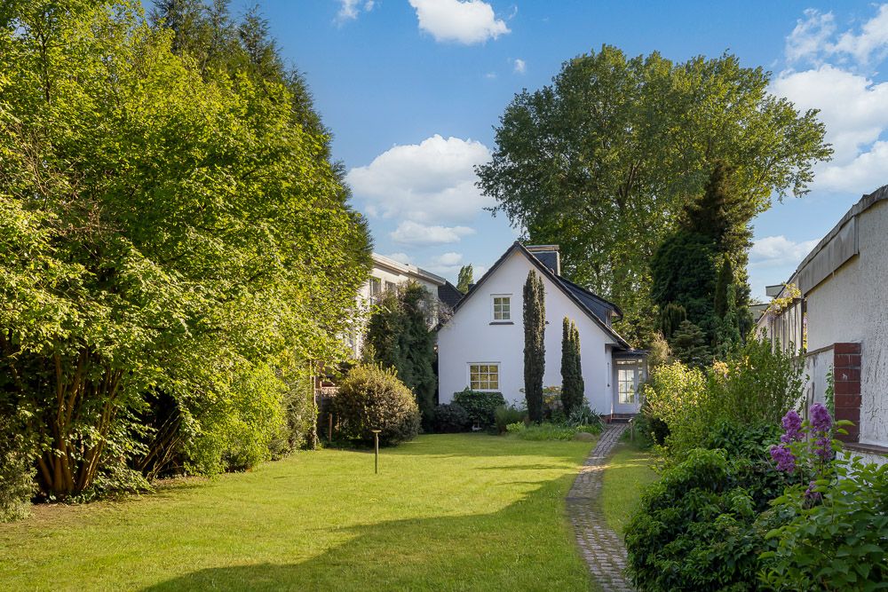 Ein weißes Einfamilienhaus mit Garten und Weg, umgeben von Bäumen und Sträuchern.