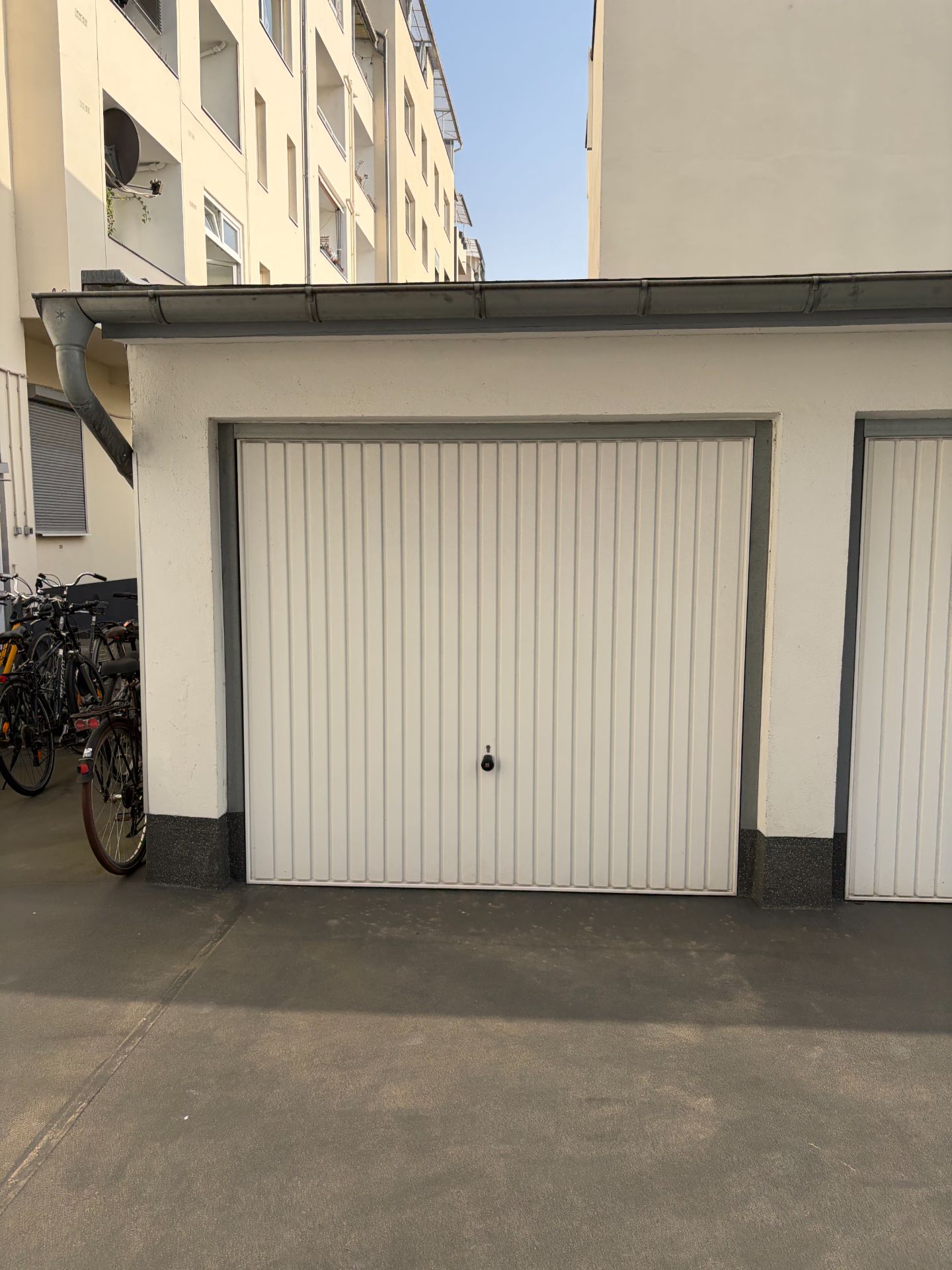 Weiße Garagentür mit Fahrrädern links, vor einem Mehrfamilienhaus.