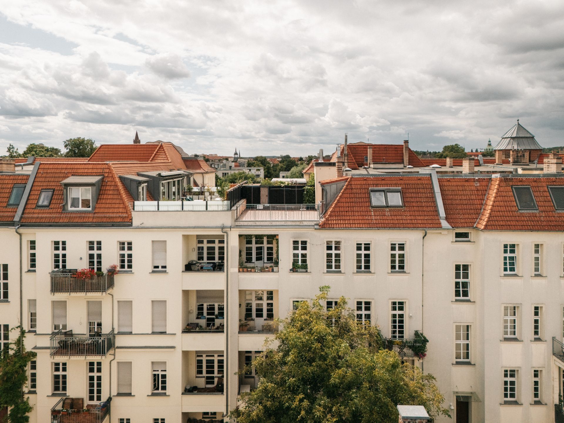 Mehrstöckiges Wohnhaus mit roten Dächern und mehreren Balkonen, davor ein Baum.