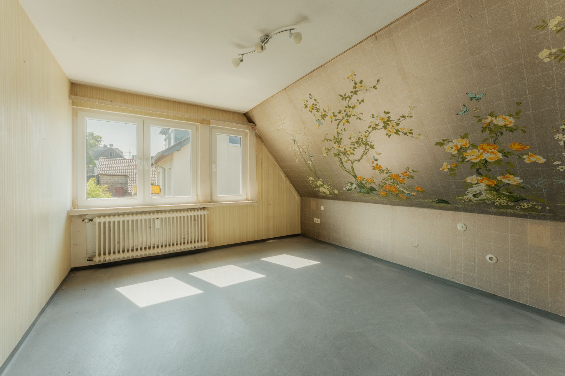 Leeres Zimmer mit großem Fenster, schräger Wand und Blumenmuster an der Wand.
