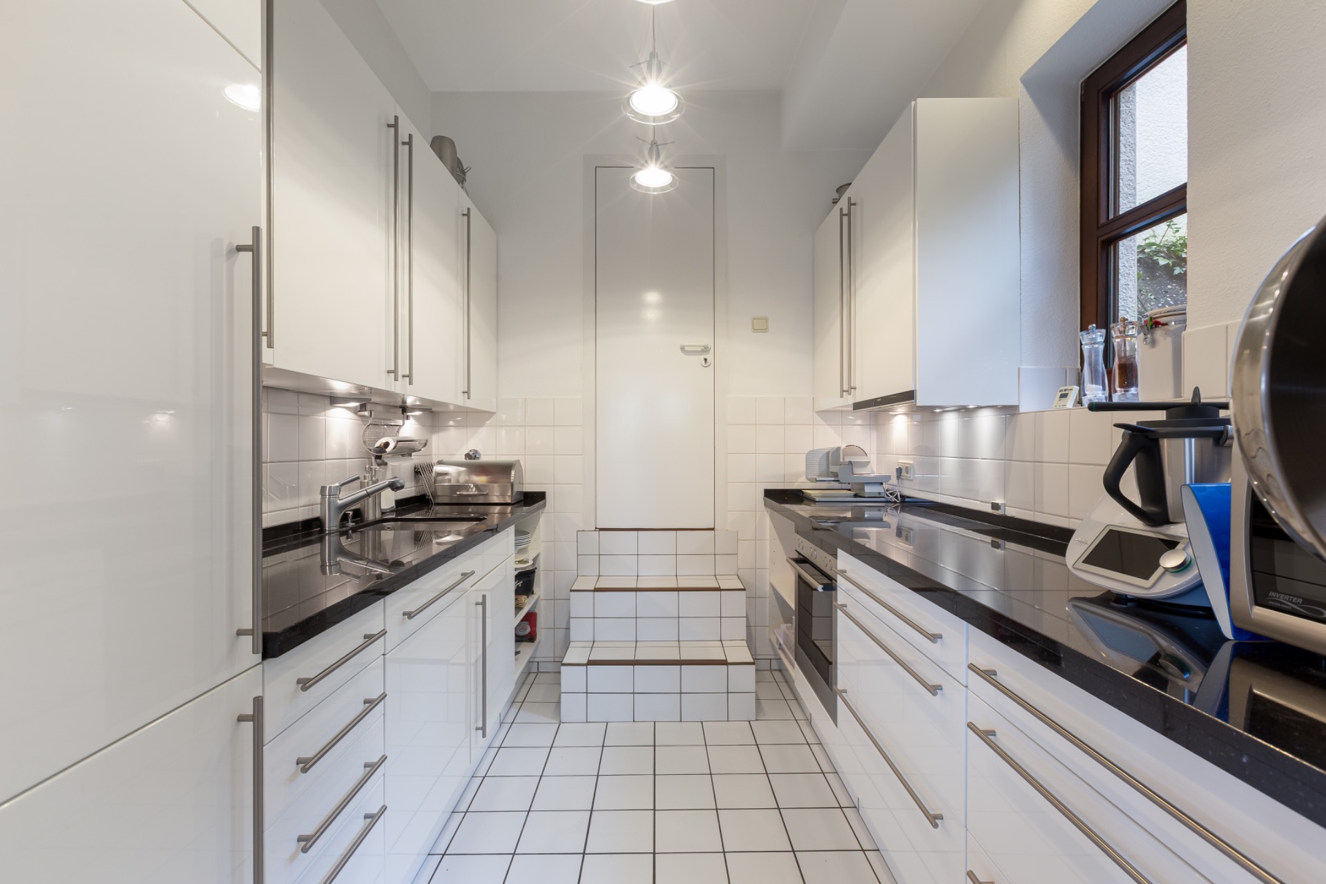 Helle, schmale Küche mit weißen Schränken, schwarzen Arbeitsplatten und Fenster rechts.