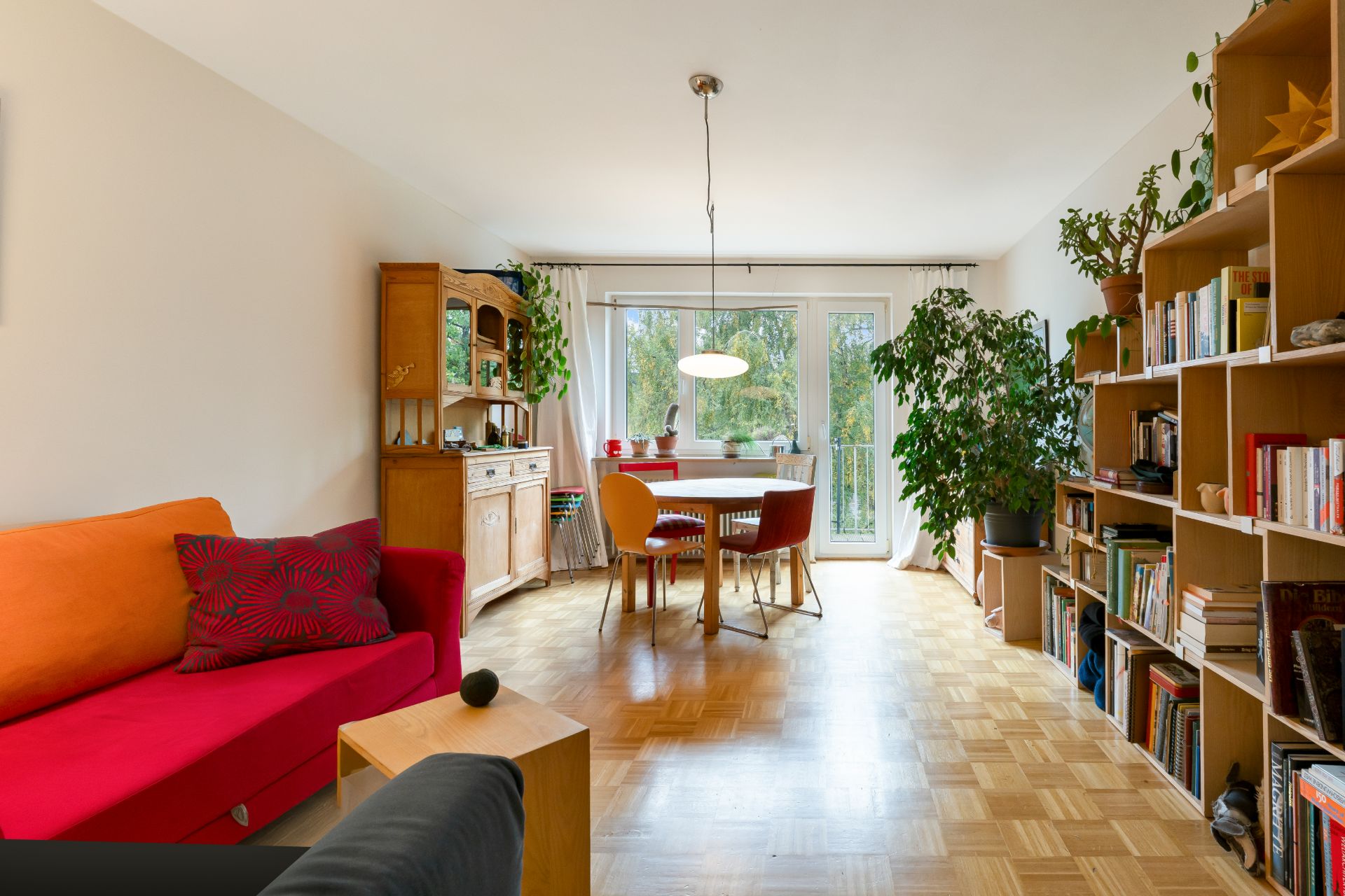 Wohnzimmer mit rotem Sofa, Esstisch, Bücherregal, Pflanzen und großen Fenstern zum Garten.