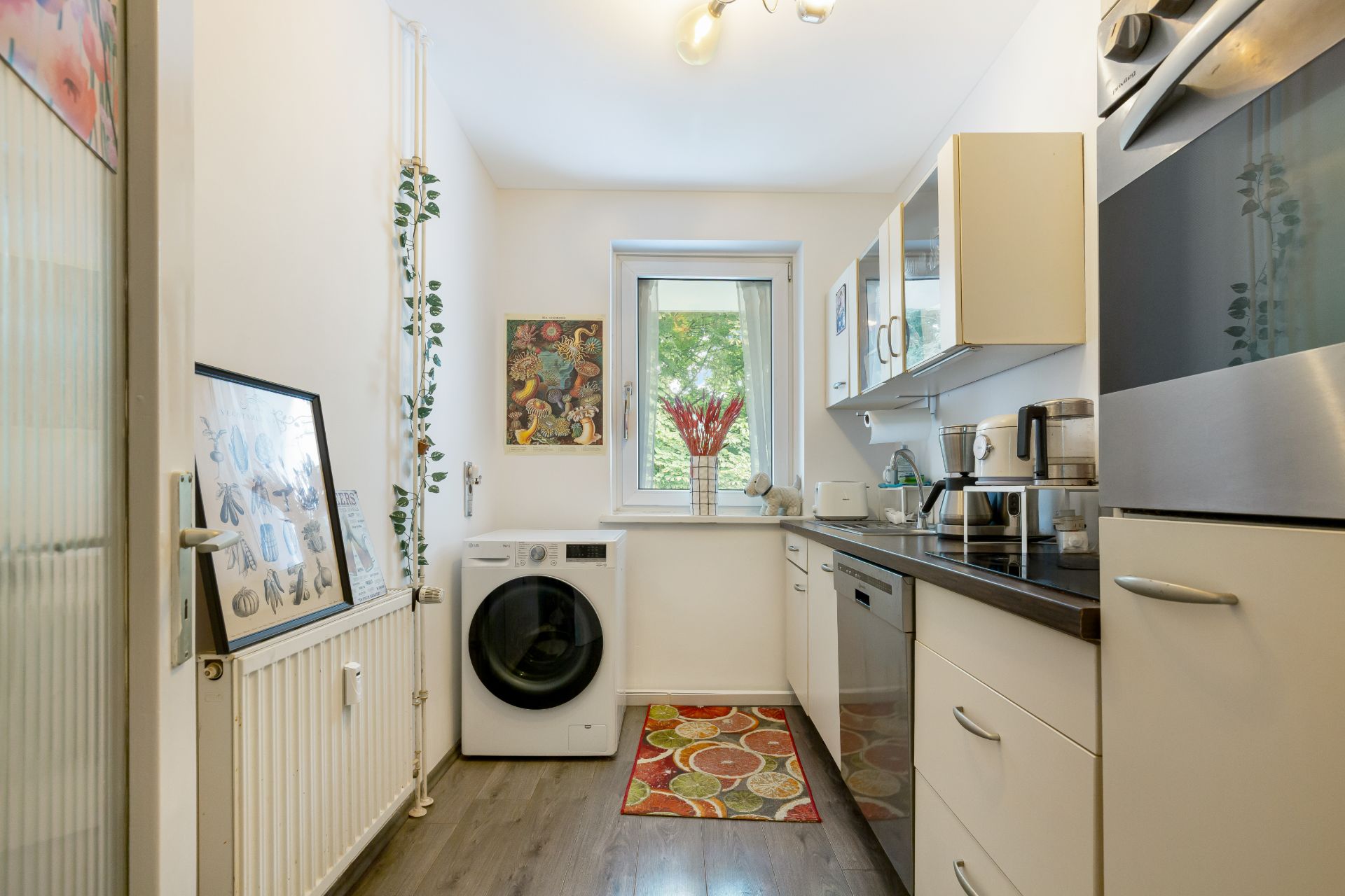 Helle Küche mit Waschmaschine, Fenster, weißen Schränken und buntem Teppich auf dem Boden.