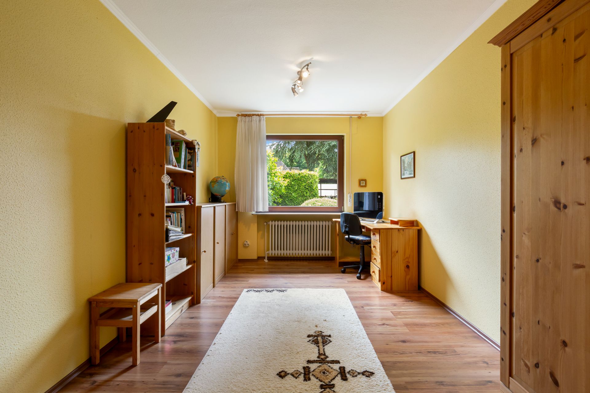 Helles Arbeitszimmer mit Holzregalen, Schreibtisch und Teppich, großes Fenster mit Blick ins Grüne.