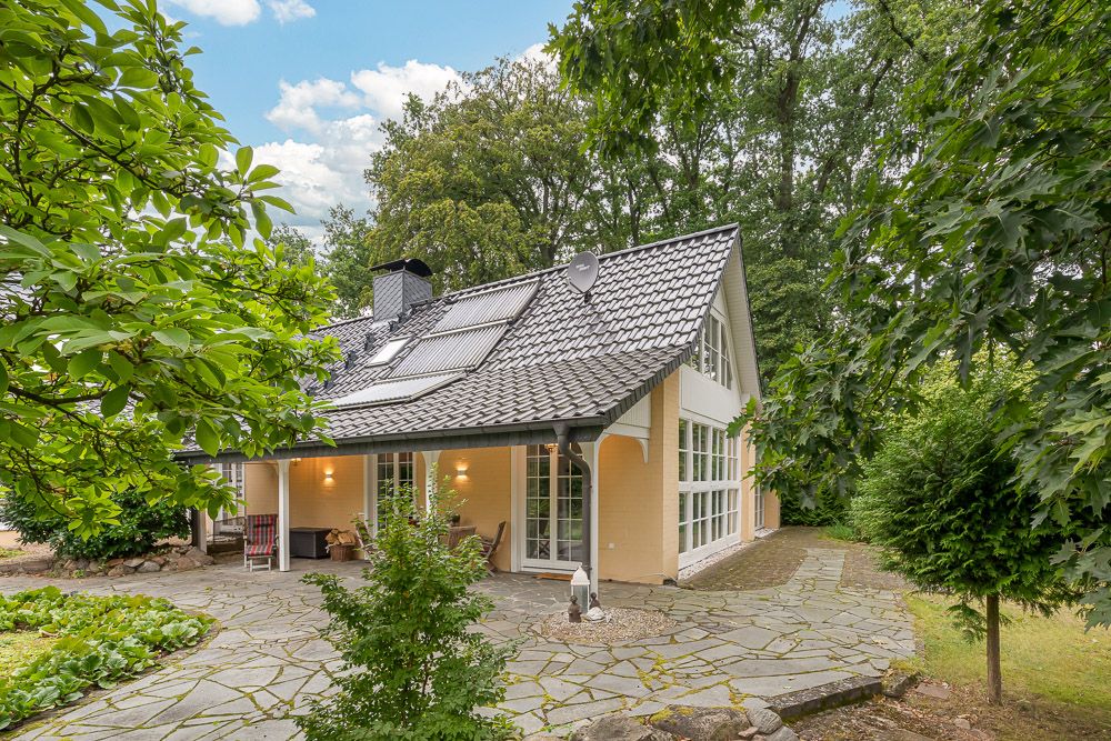 Modernes Einfamilienhaus mit großem Fenster, Terrasse und Garten, umgeben von Bäumen.