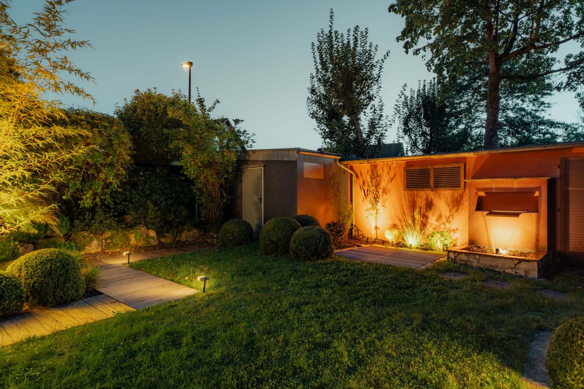Beleuchteter Garten mit Rasen, Holzboden, Büschen und einem kleinen orangefarbenen Gebäude bei Abenddämmerung.